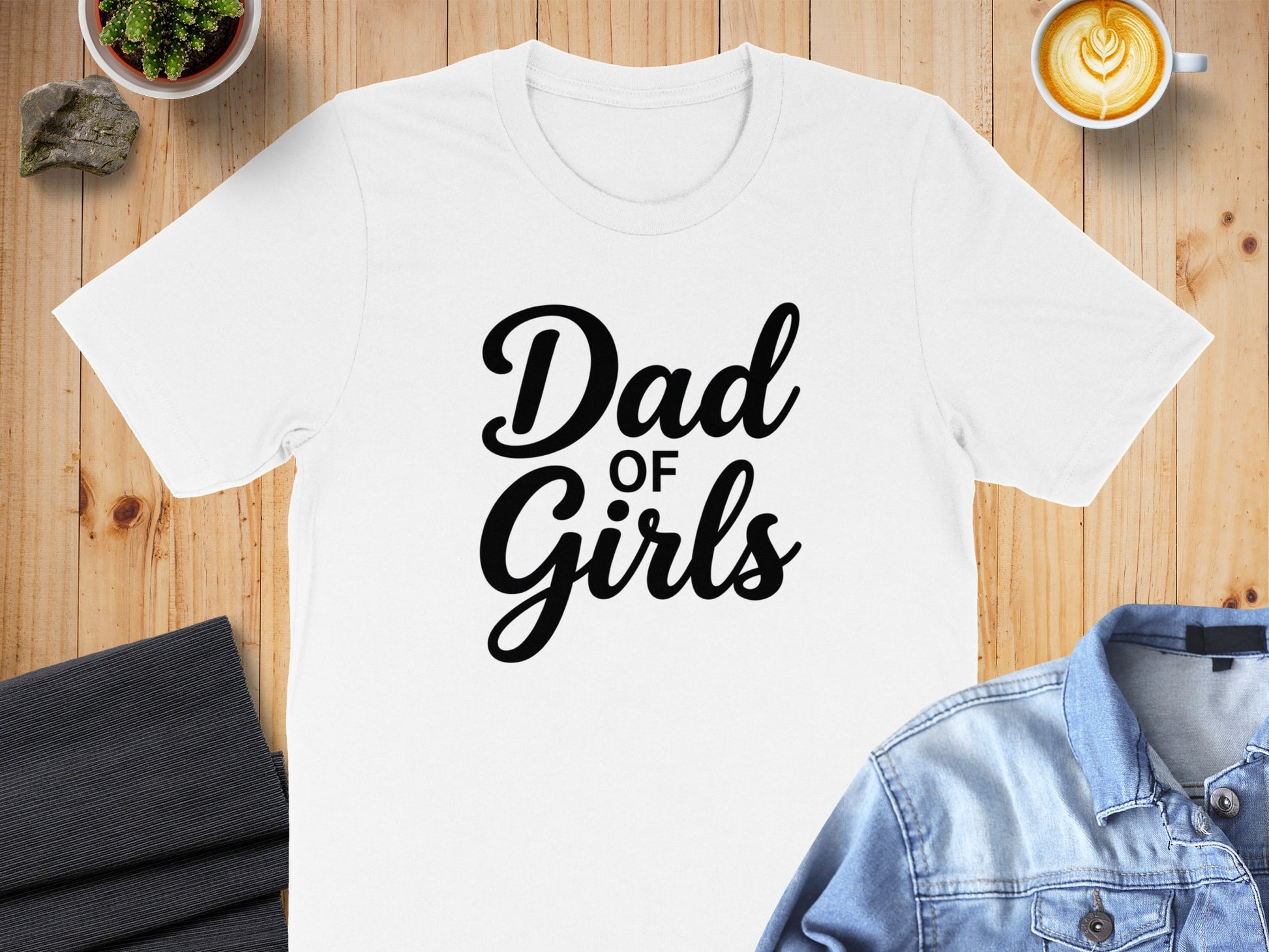 White t-shirt with "Dad of Girls" text, flat lay with denim jacket and coffee.